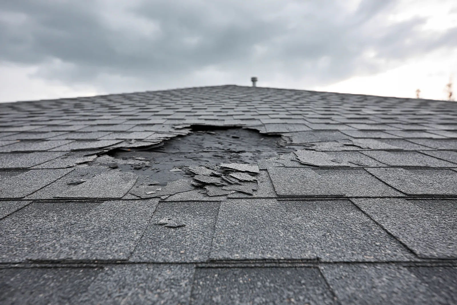 Beneath a stormy sky torn shingles reveal a hole in the roof